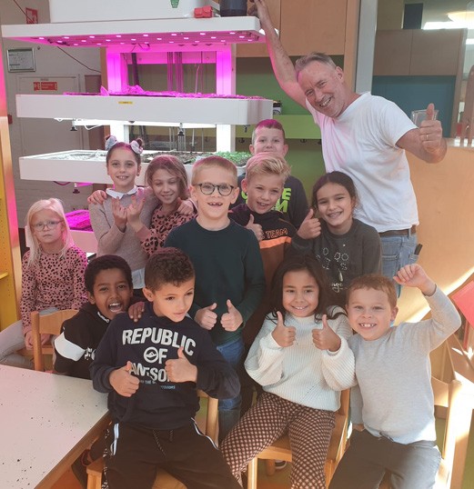 Groep blije kinderen en leerkracht poseren bij een verticale tuin belicht met roze LED-licht.