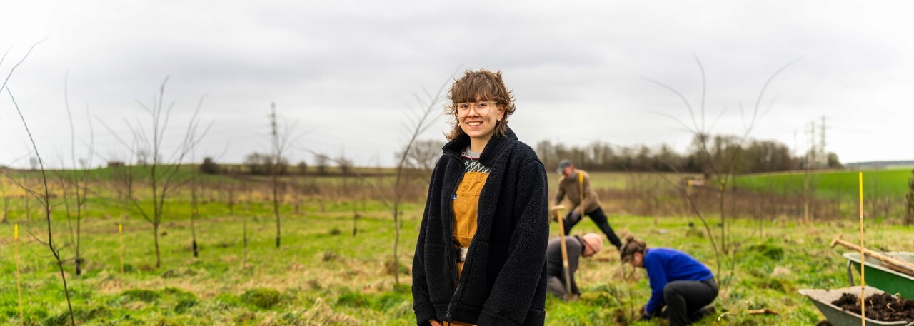 Een persoon glimlacht naar de camera; anderen planten bomen in een veld.