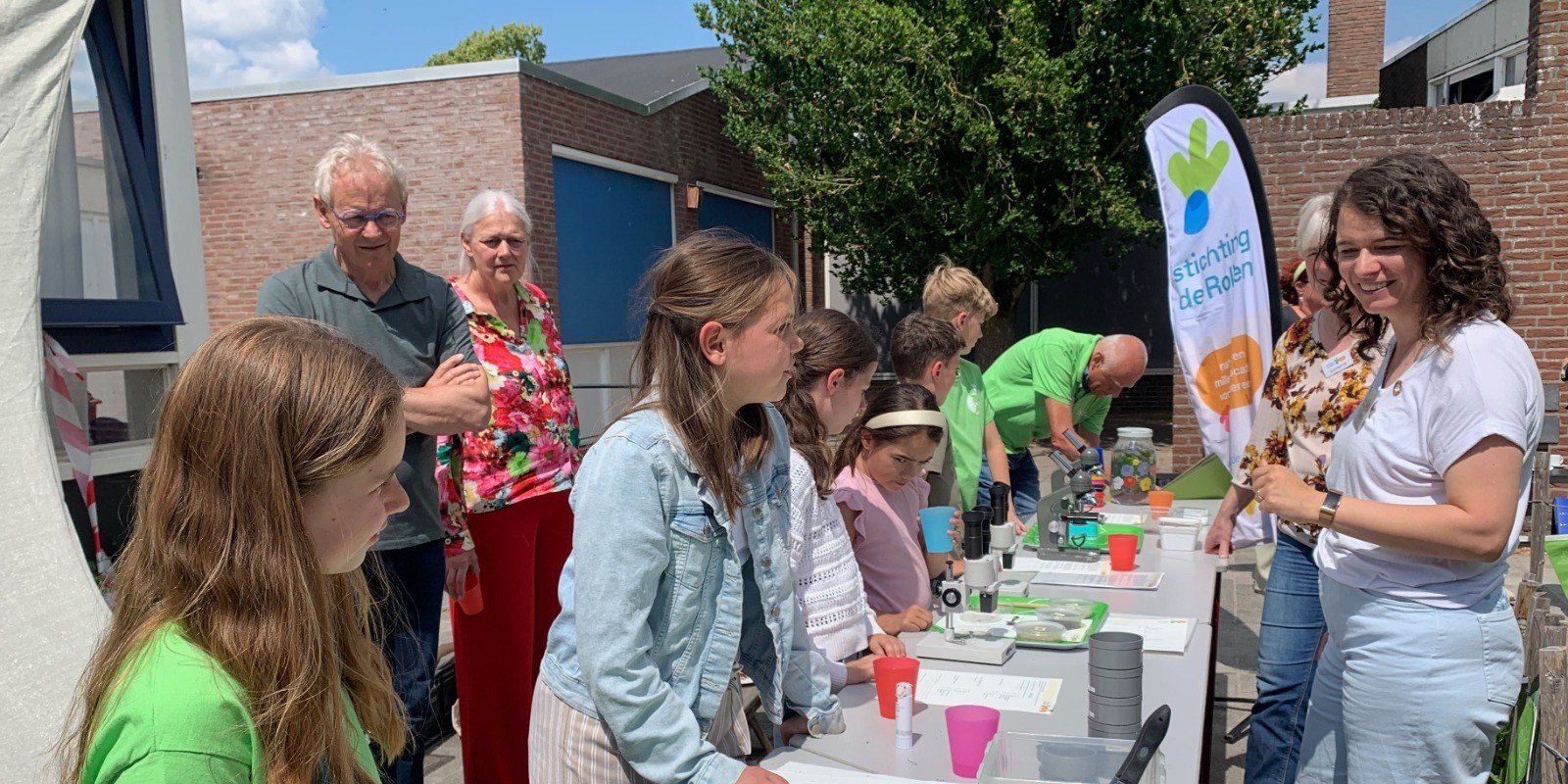 Groep mensen bezig met wetenschappelijke experimenten aan tafels buiten. Banner met logo op achtergrond.