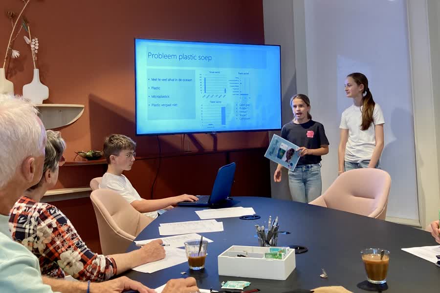 Jongeren geven presentatie over plasticsoep voor een kleine groep volwassenen in een vergaderruimte.