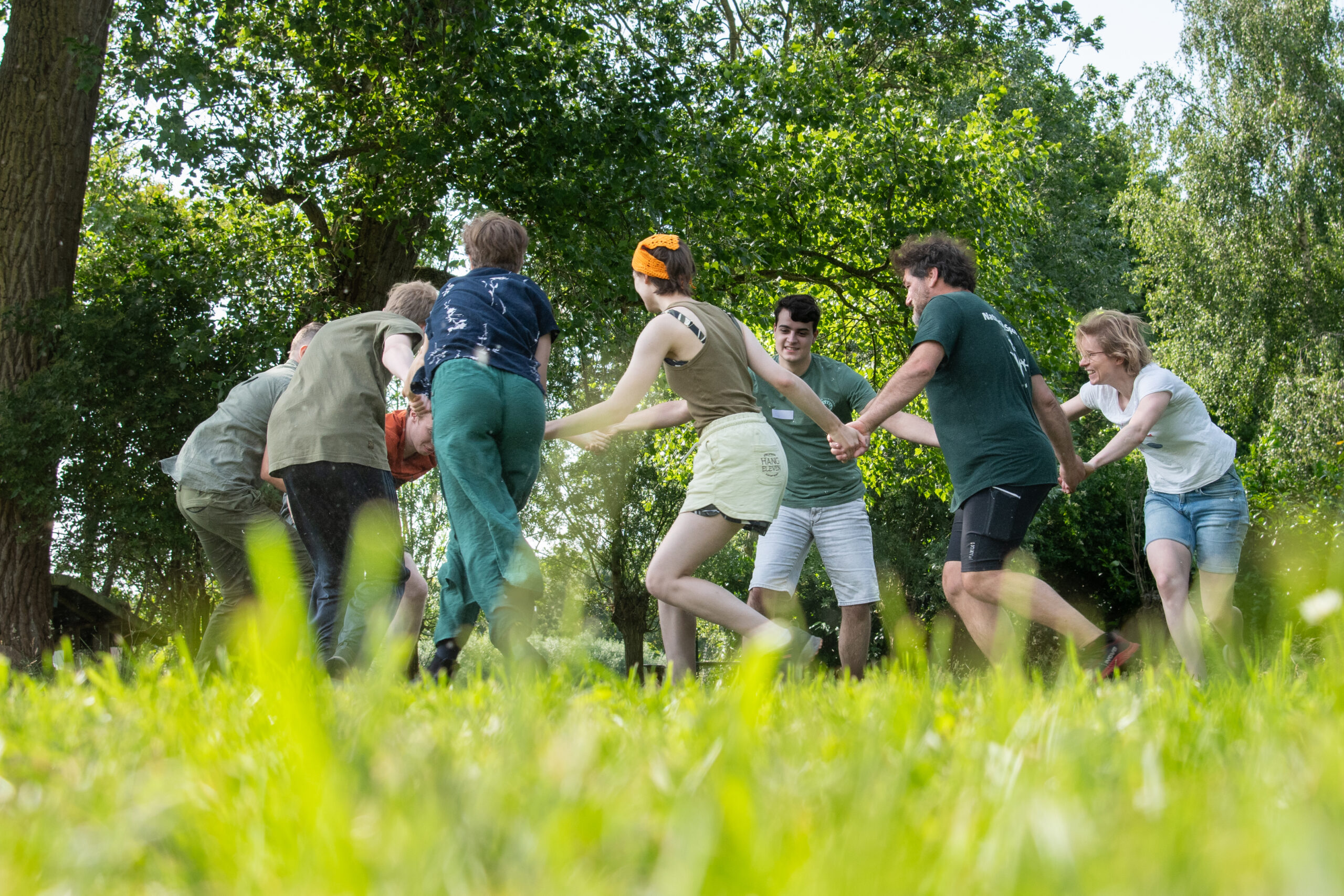 Een groep mensen houdt elkaars handen vast in een kring op een grasveld, omringd door bomen.