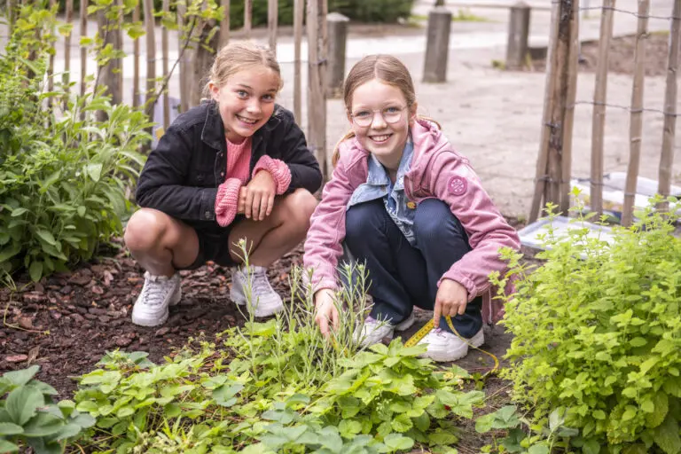 Twee lachende kinderen verzorgen een plant in een tuin. Ze zitten gehurkt tussen groen en houten hekjes.