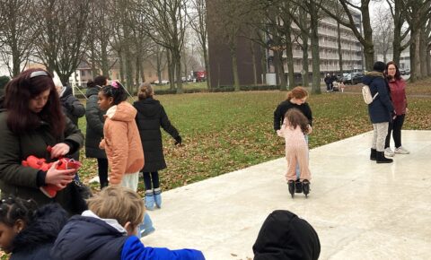 Kinderen en volwassenen schaatsen op een buitenbaan, omringd door bomen en gebouwen.