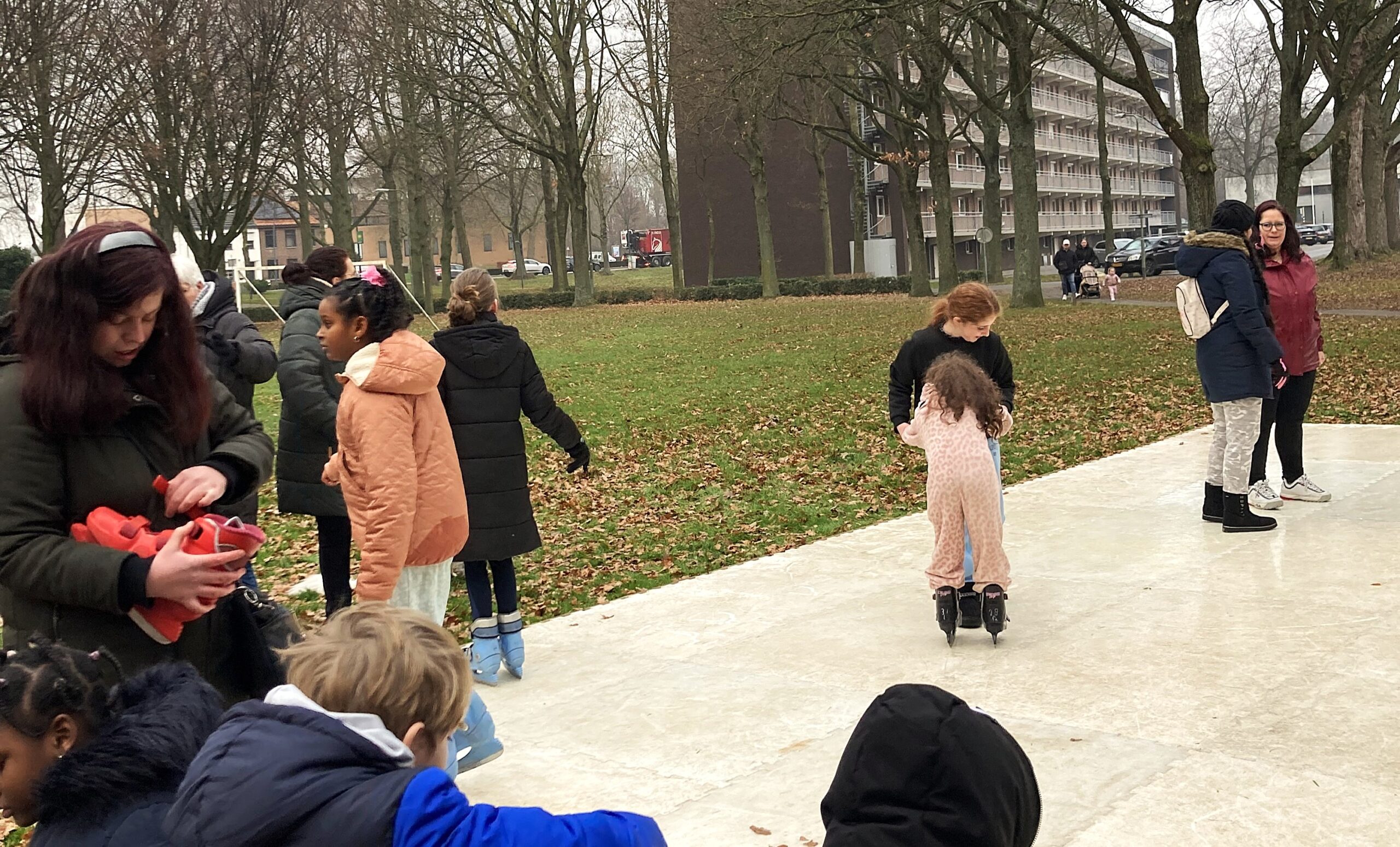 Kinderen en volwassenen schaatsen op een buitenbaan, omringd door bomen en gebouwen.