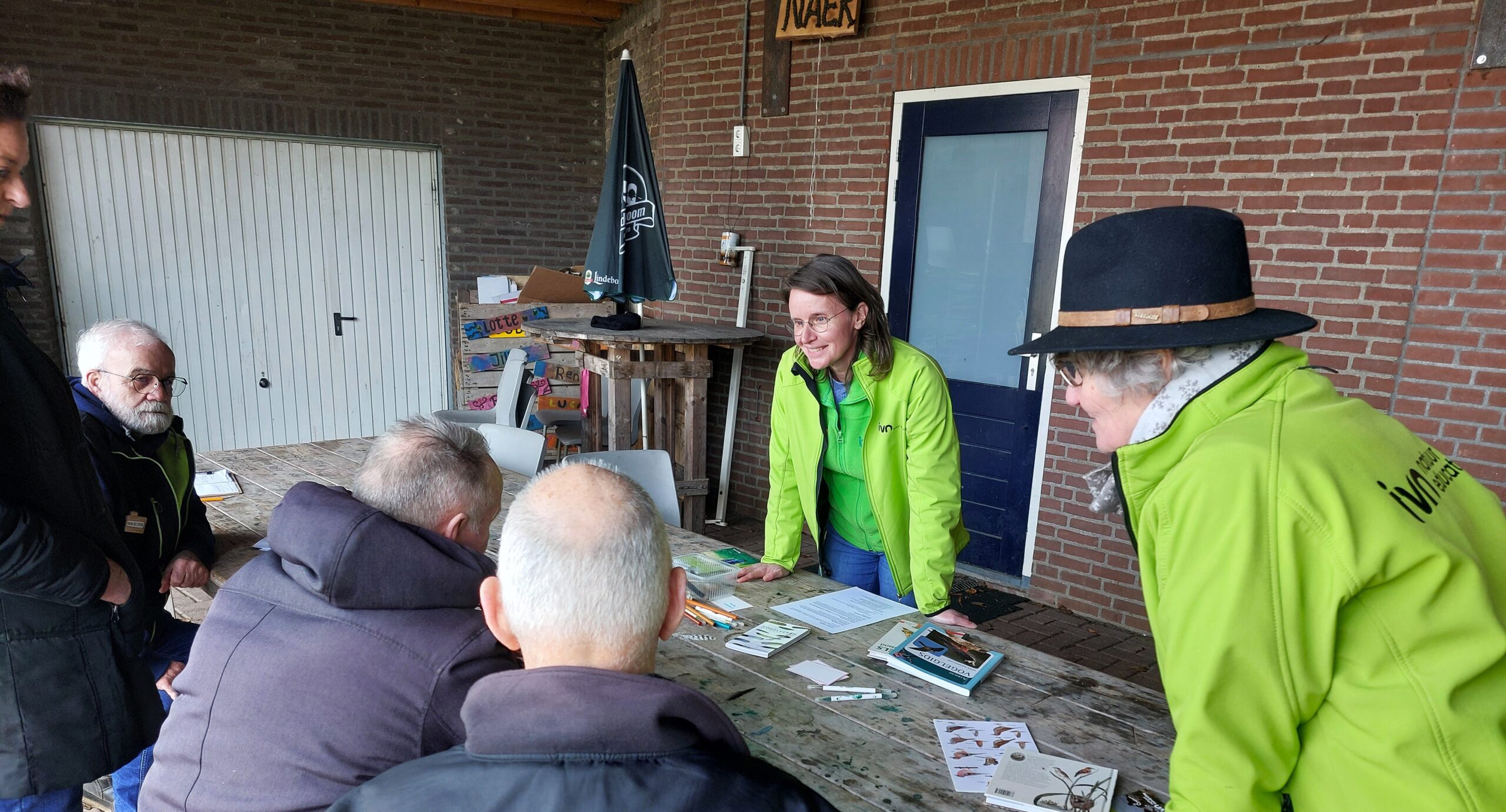Een vrouw in groene jas geeft uitleg aan zittende mensen aan een tafel met folders en boeken.