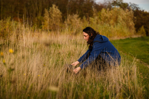Persoon hurkt in hoog gras met een blauwe trui in een herfstlandschap.