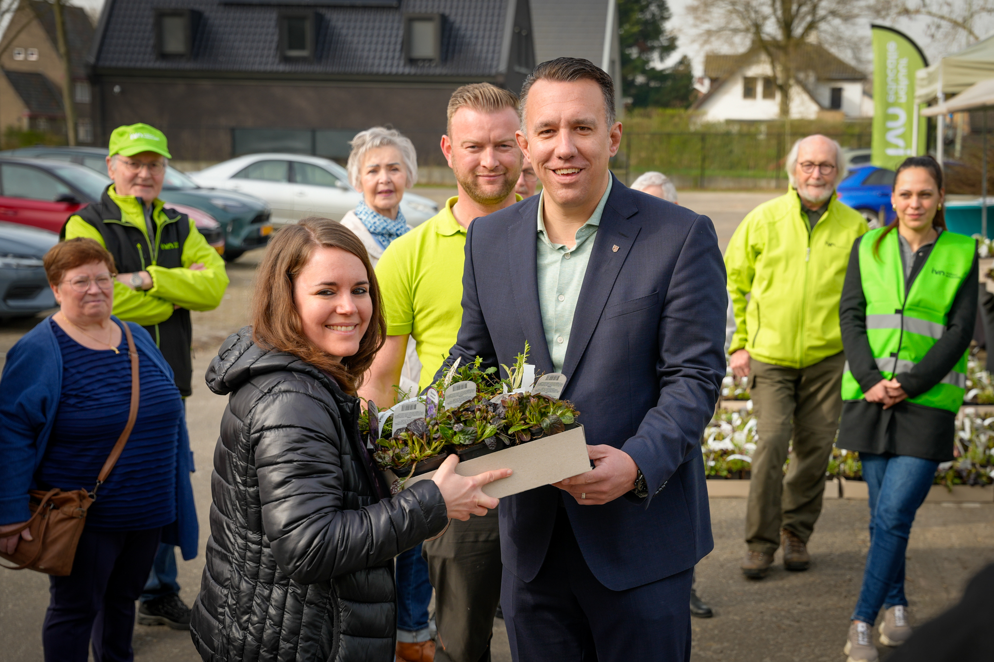 Man en vrouw overhandigen plantenbak, omringd door groep mensen buiten bij een evenement.