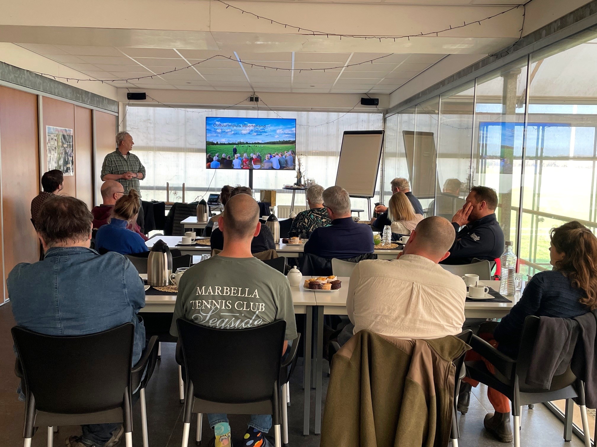 Mensen luisteren naar een presentatie in een vergaderruimte met een scherm en flip-over.