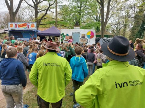 Groep kinderen in openluchtbijeenkomst voor theaterwagen, begeleid door IVN natuur educatie.