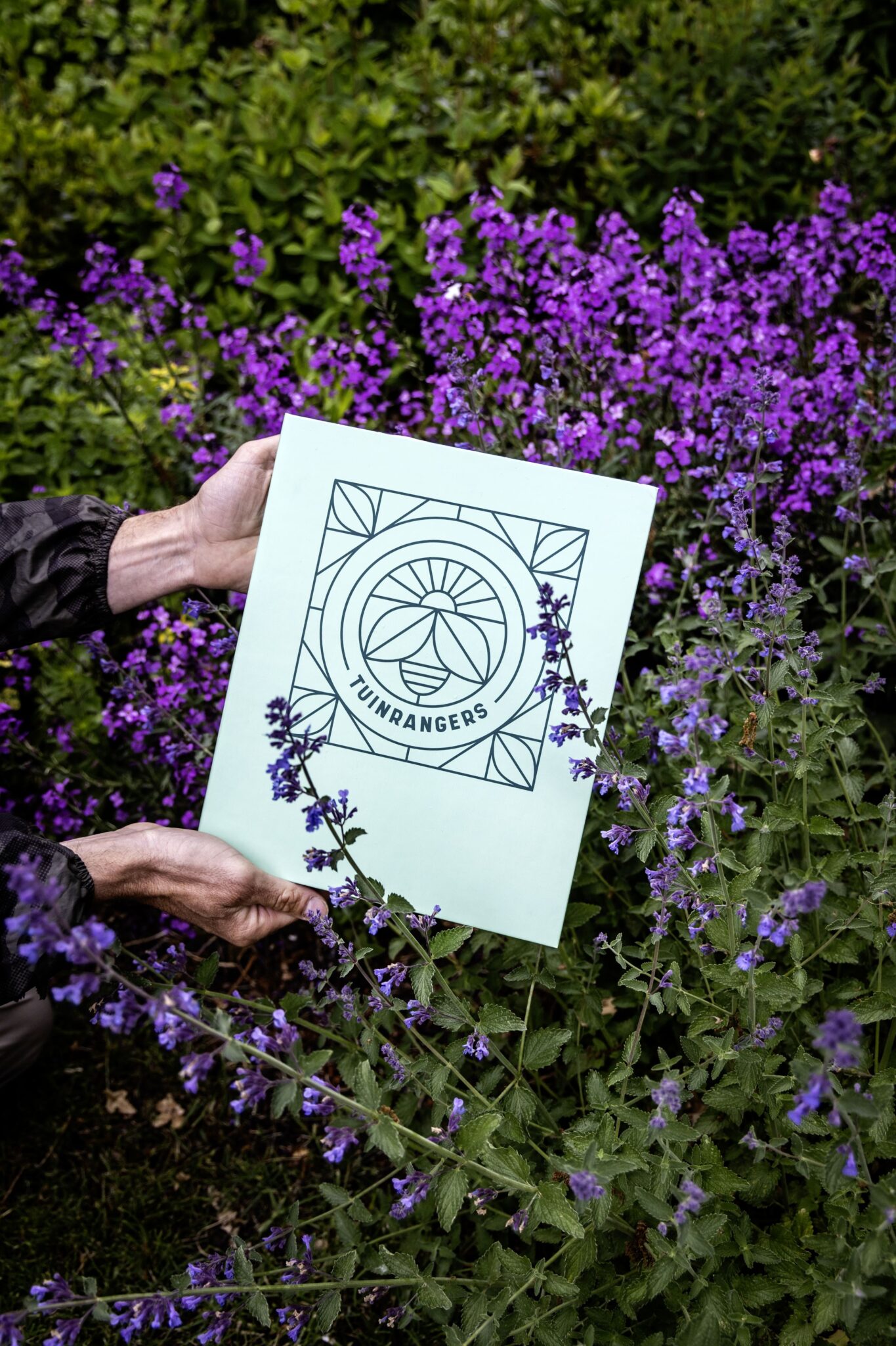 Twee handen houden een Tuinrangers-bord vast te midden van paarse bloemen en groen.