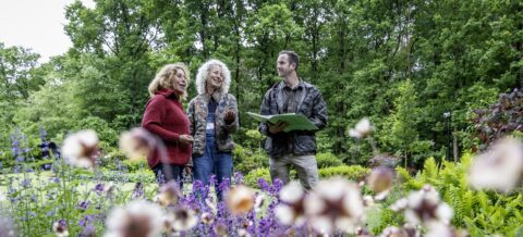 Drie mensen praten in een groen park tussen bloeiende bloemen.