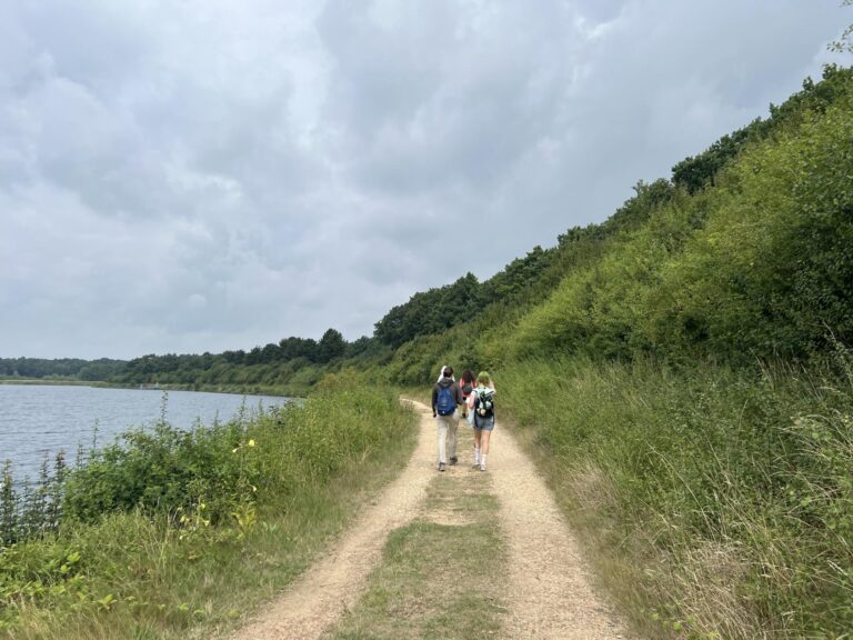 Twee mensen wandelen op een pad langs een meer met bewolkte lucht en groene heuvels.