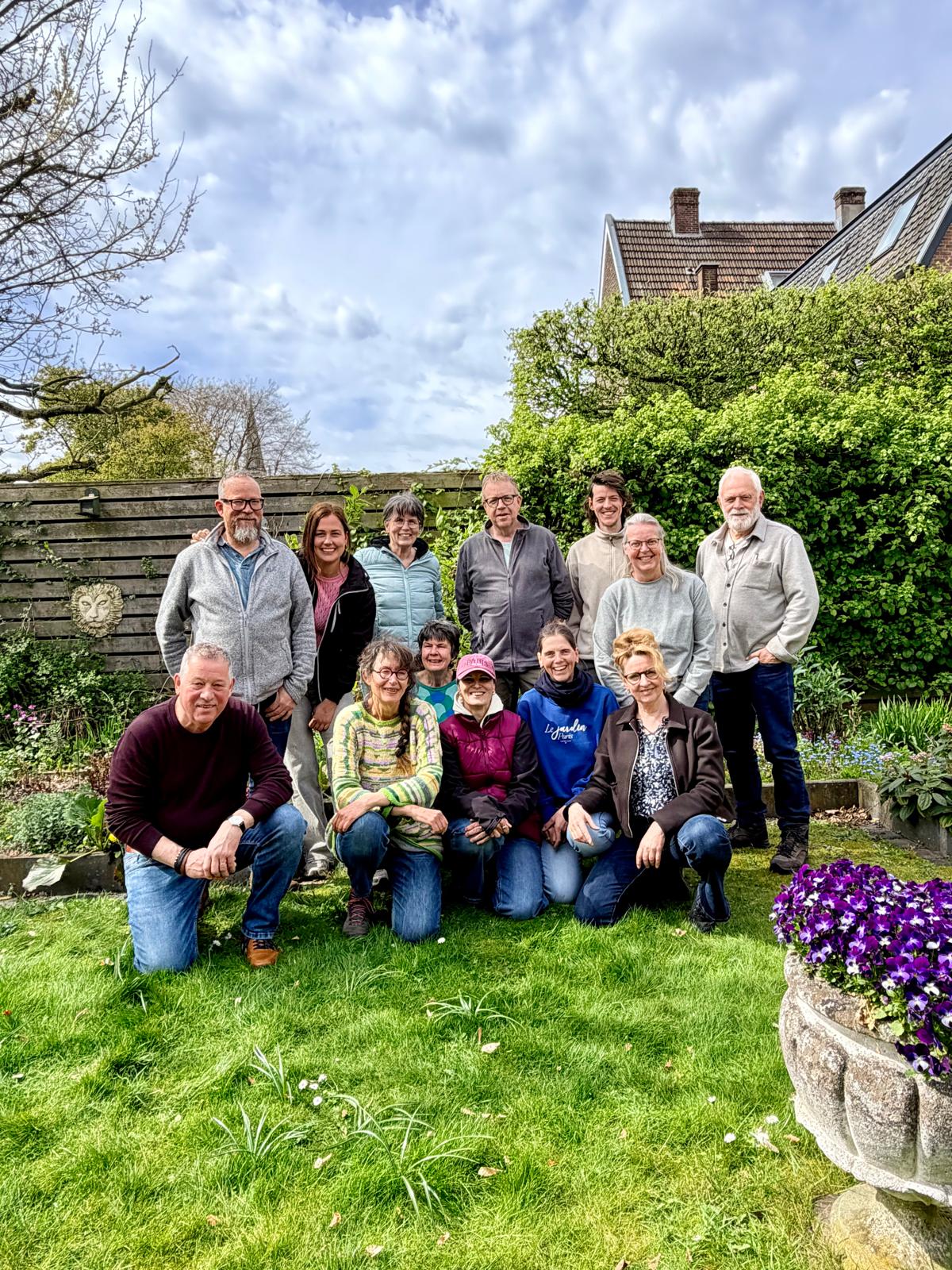 Groep mensen poseert in een groene tuin voor een houten schutting met bloeiende planten.