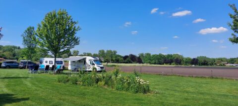 Kampeerplek met camper, caravans, tuinstoelen en bomen op een zonnig veld.