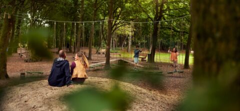 Twee volwassenen zitten in bos bij speelpark, twee kinderen spelen op touwbruggen.