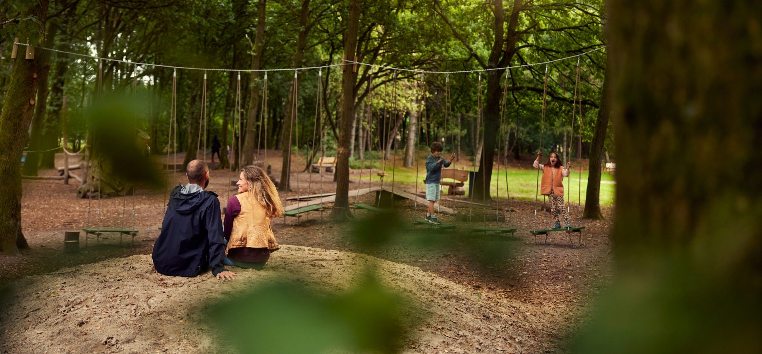 Twee volwassenen zitten in bos bij speelpark, twee kinderen spelen op touwbruggen.