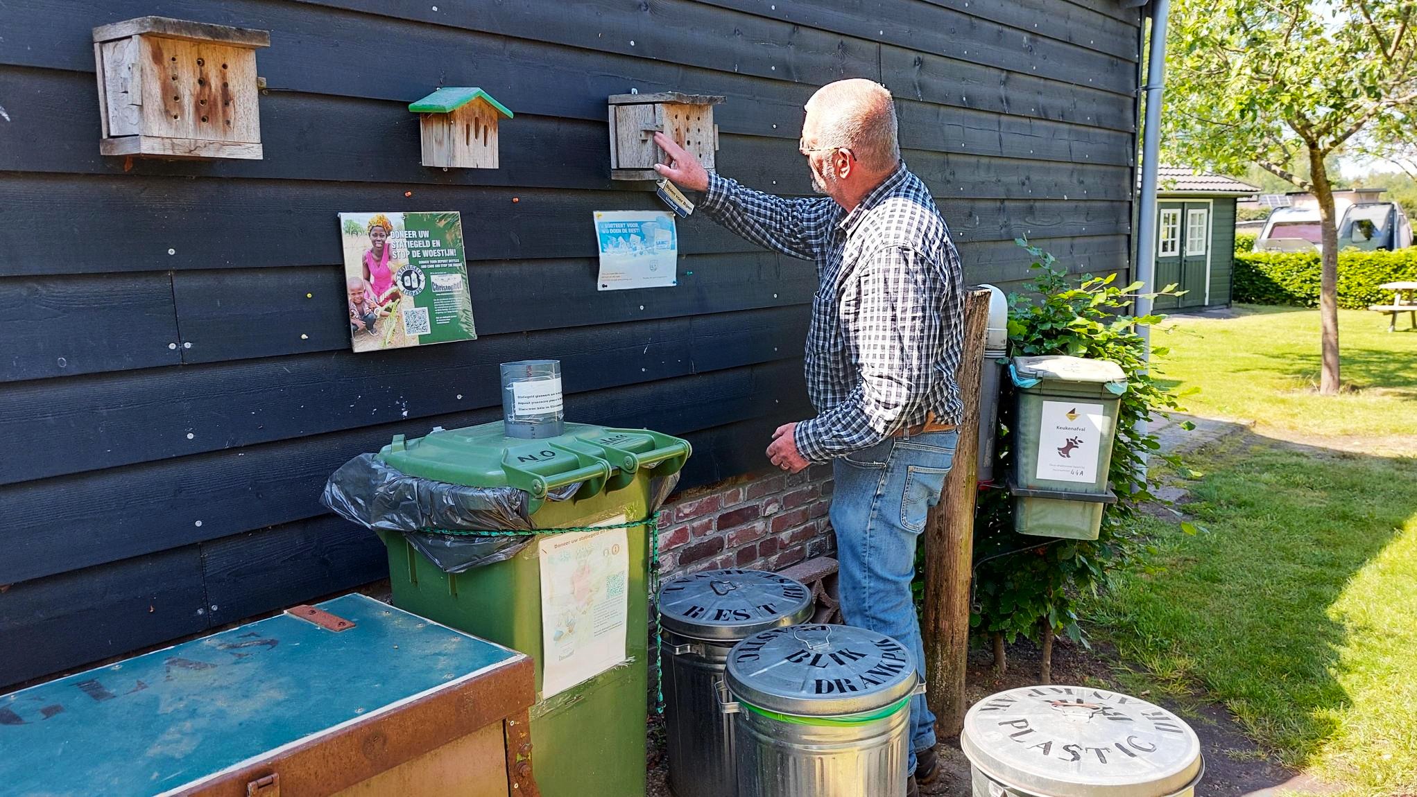 Man bij houten wand met vogelhuisjes, afvalbakken en posters, in een tuinomgeving.