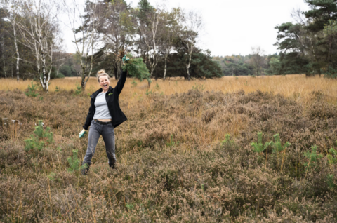 Vrouw juicht in een open veld, houdt een dennentak omhoog, omringd door bomen en gras.
