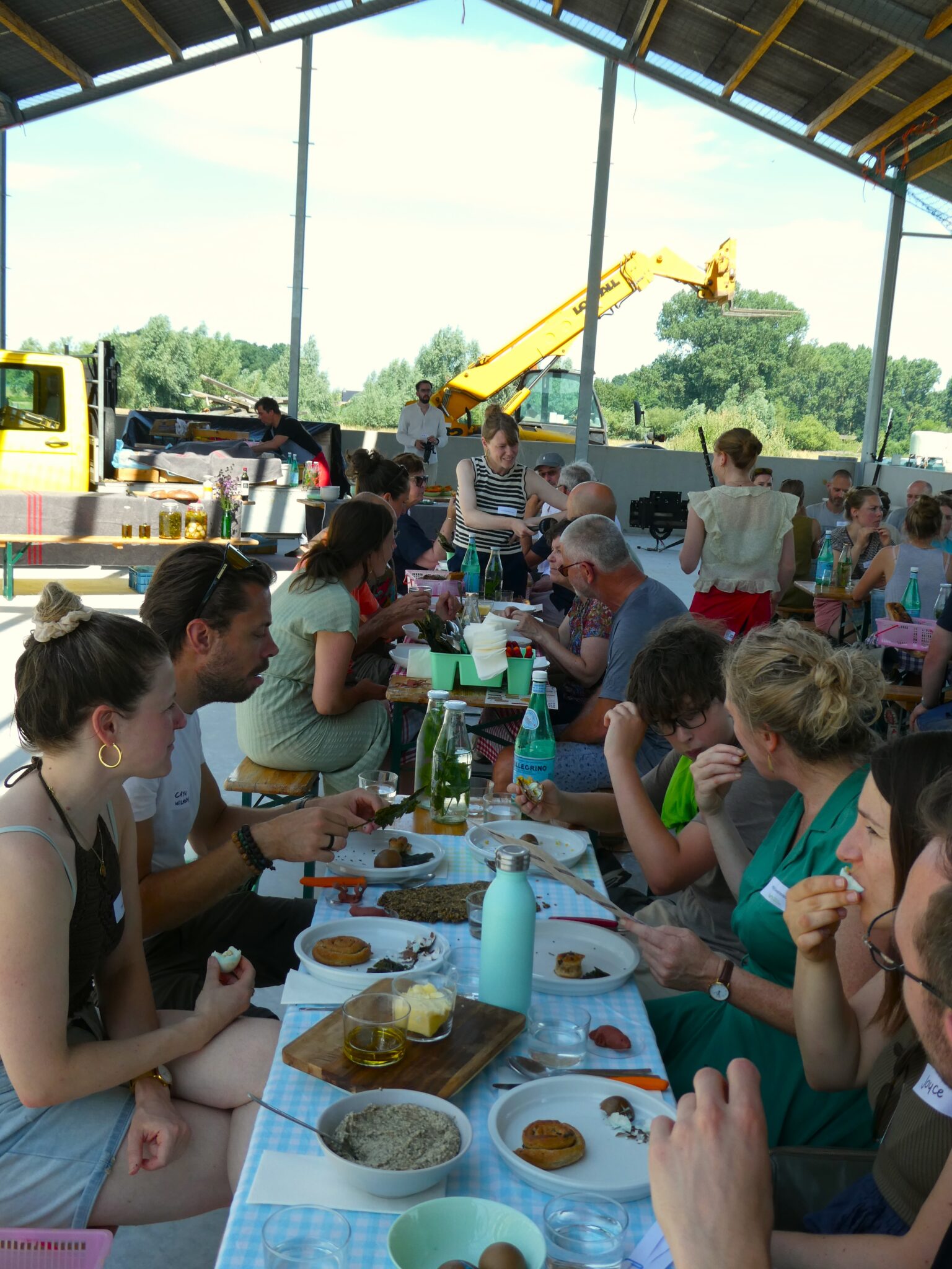 Mensen eten en praten aan een lange tafel in een openlucht loods, met bouwmachines op de achtergrond.