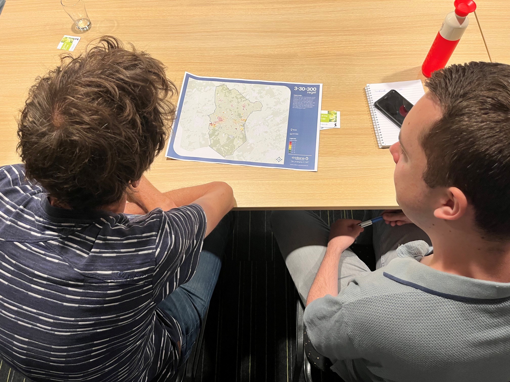 Twee mannen bekijken een kaart op tafel met een notitieblok, mobiele telefoon en waterfles.