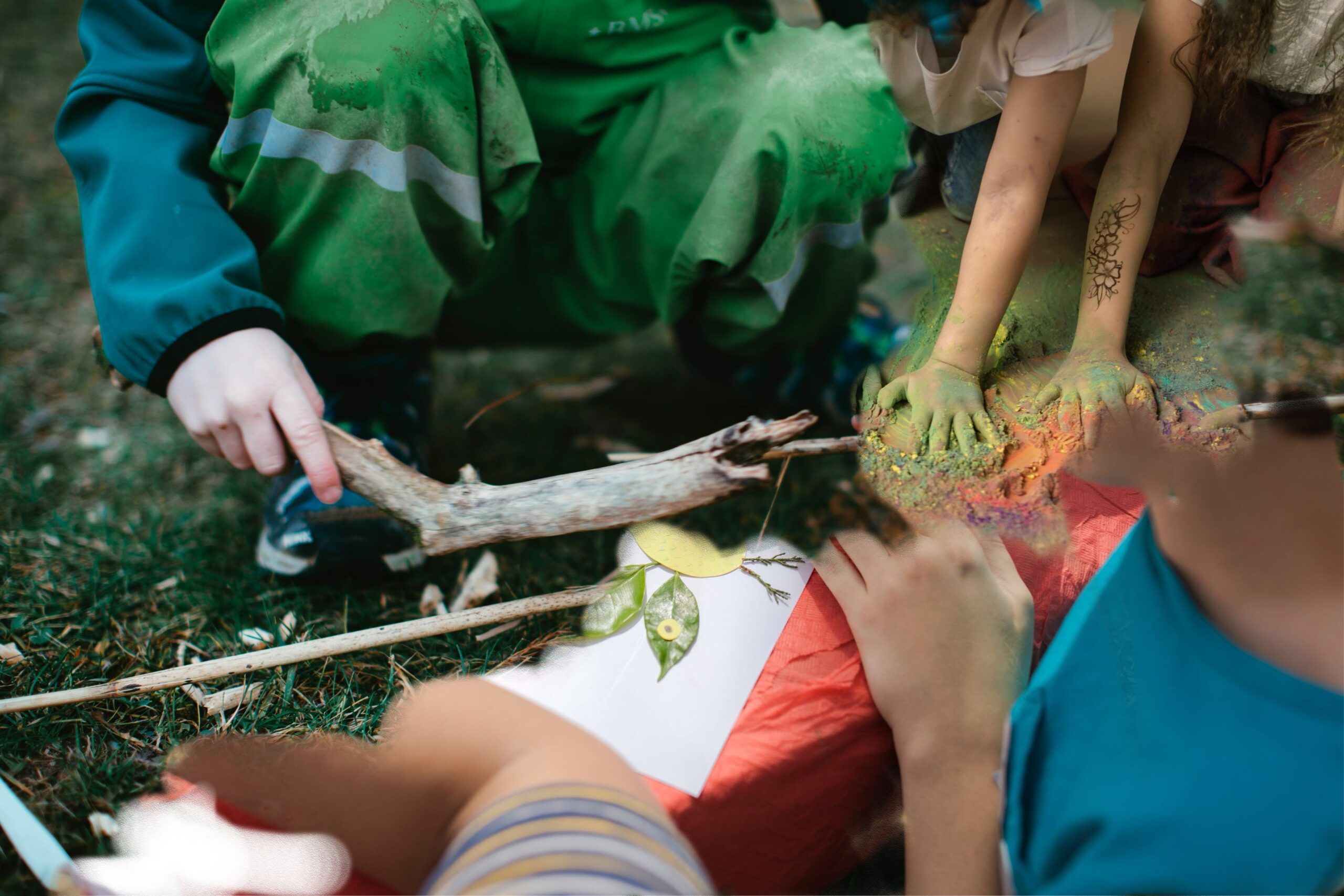 Kinderen spelen buiten met takken en kleuren, groene en kleurrijke handen zichtbaar.