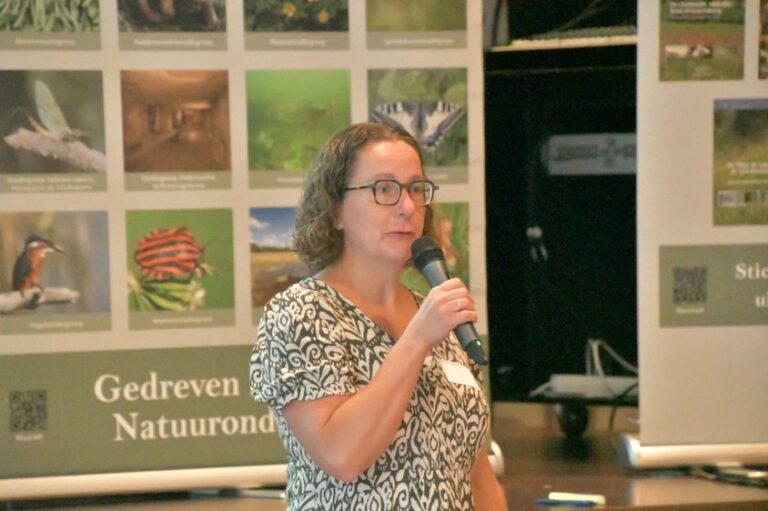 Vrouw spreekt in microfoon bij poster met natuurafbeeldingen en tekst 