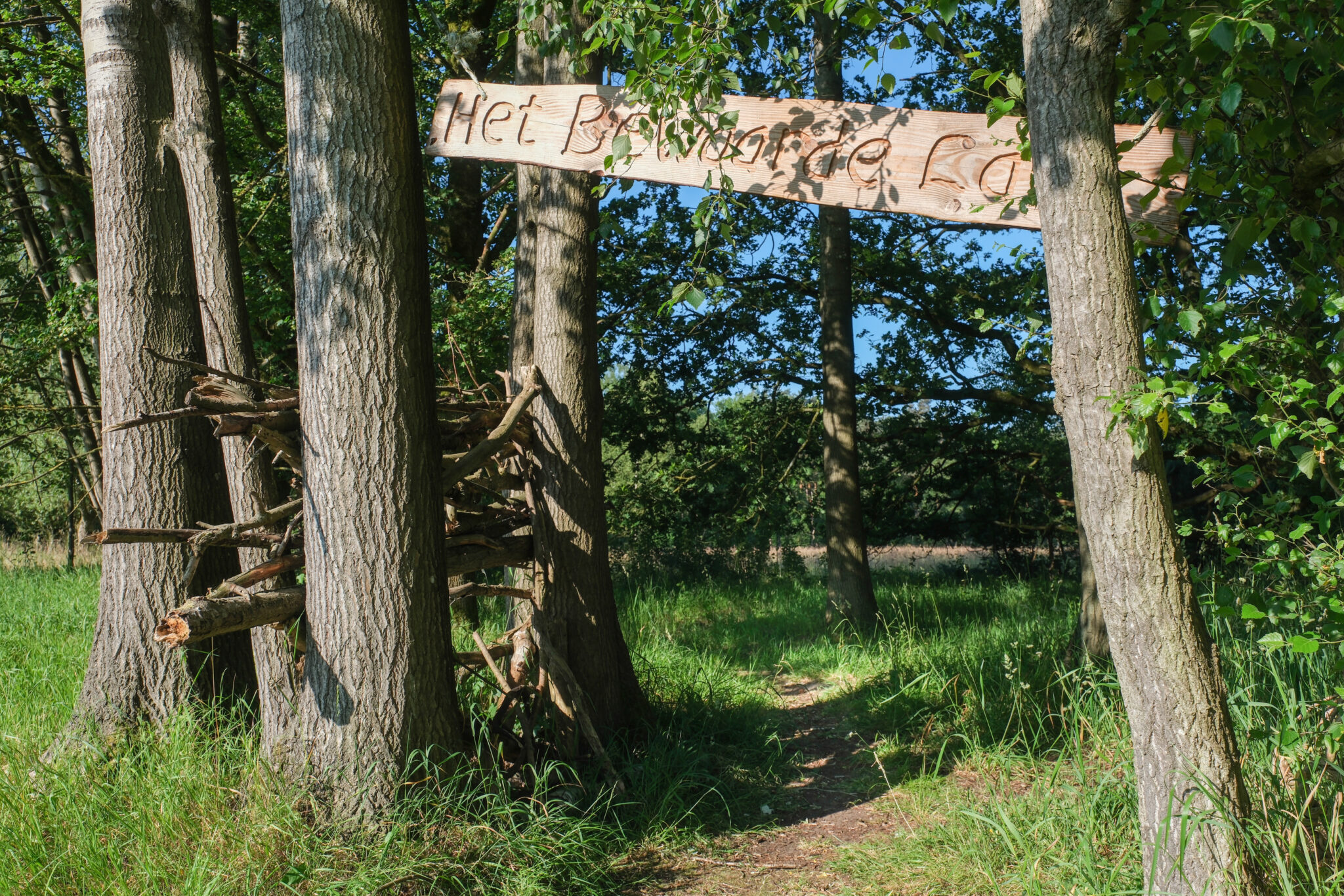 Houten bord met "Het Bewaarde Land" boven een bospaadje tussen bomen.