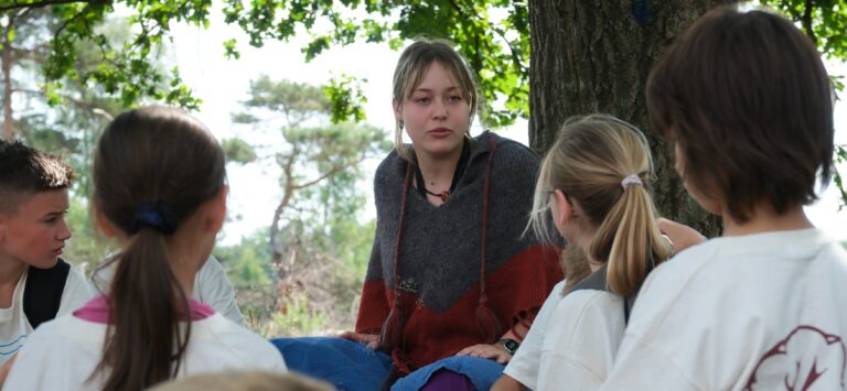 Vrouw praat met kinderen onder een boom in een bosrijke omgeving.