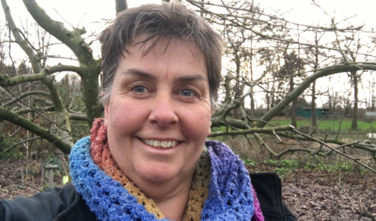 Vrouw in veelkleurige sjaal glimlacht buiten voor kale bomen.