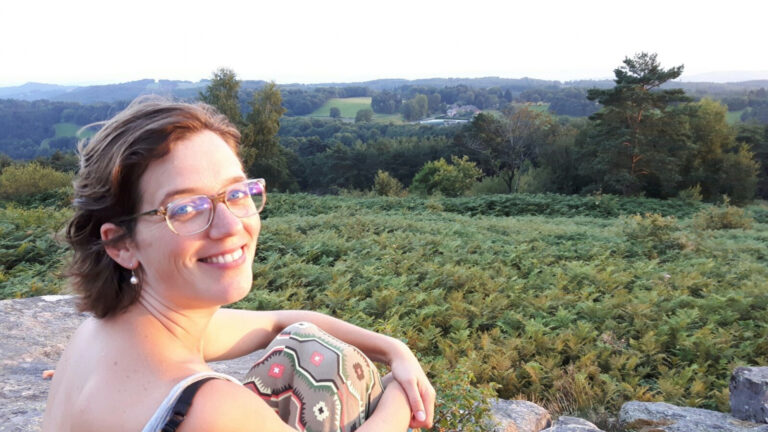 Vrouw glimlacht naar de camera met een groene heuvelachtige achtergrond bij zonsondergang.