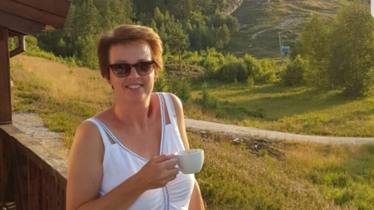 Vrouw glimlacht met een kleine kop koffie, buiten met groene heuvels op de achtergrond.