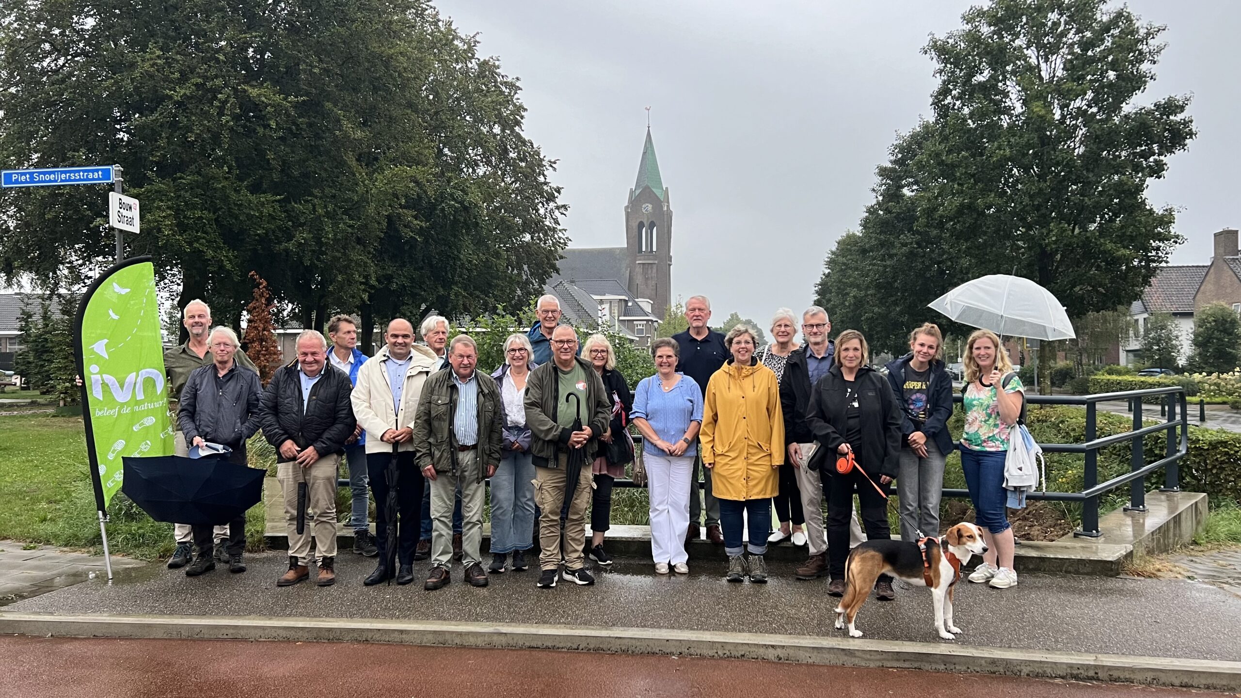 Groep mensen poseert buiten met een IVN-vlag en een kerk op de achtergrond, naast straatnaamborden.