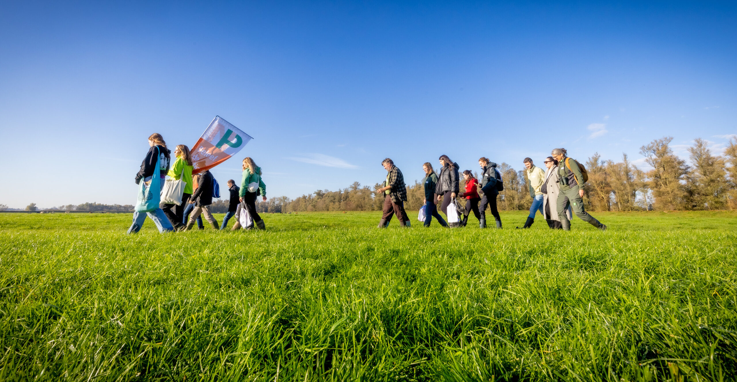 Groep mensen wandelt over een groene weide onder een heldere blauwe hemel, met een vlag.