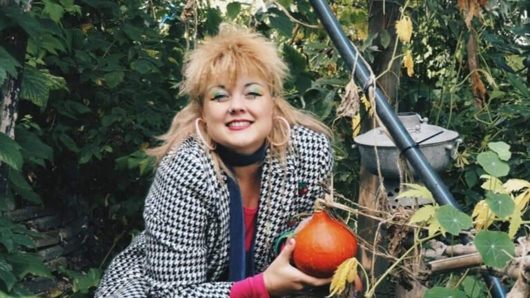 Vrouw met oranje pompoen in dichte, groene tuin met grote oorbellen en geruite jas.