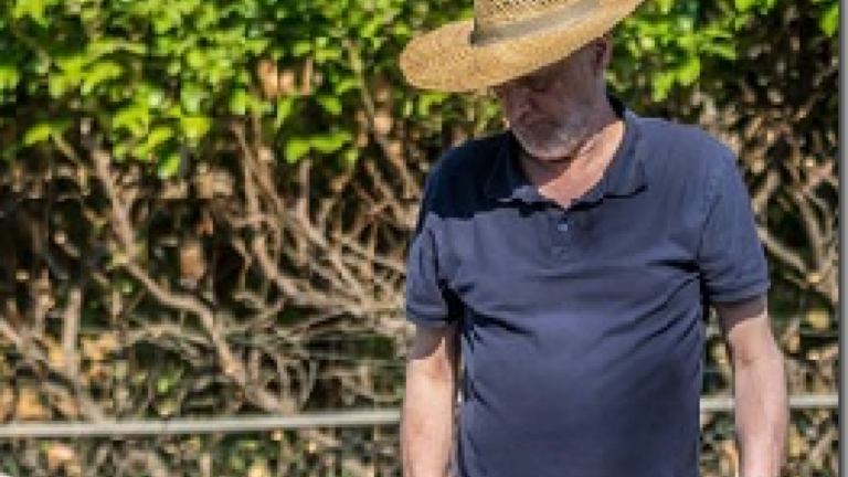 Een man met een strohoed en donker shirt staat voor groene struiken.