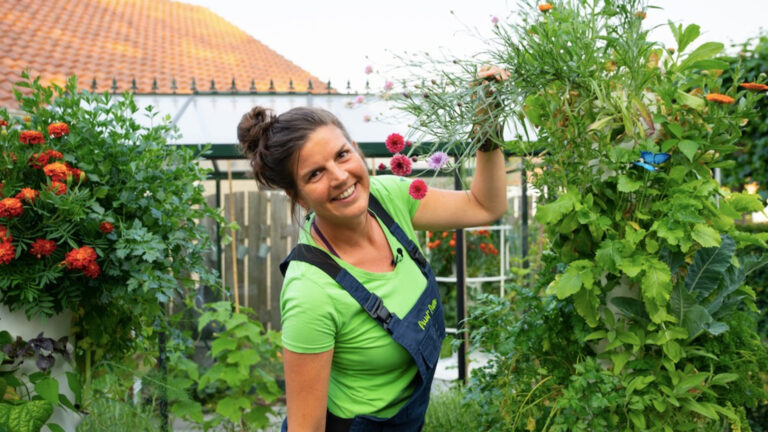 Vrouw in groene tuin werkt met bloemen, lachend naar de camera.