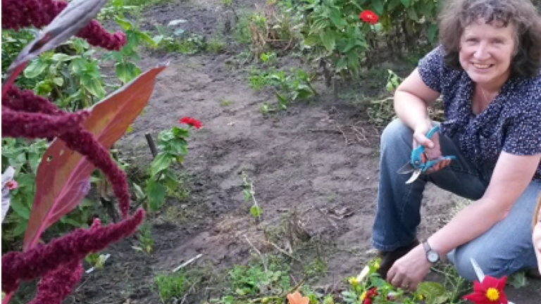 Vrouw knielt in tuin met bloemen, glimlachend, houdt snoeischaar vast.