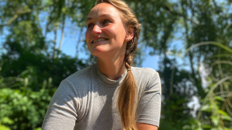 Vrouw glimlacht buiten in de zon, met groene bomen in de achtergrond.