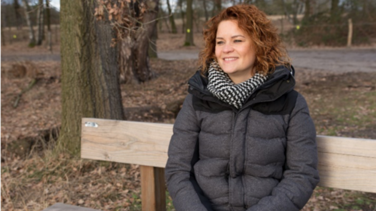 Vrouw met krullend haar glimlacht op houten bank in bos, draagt warme jas en sjaal.