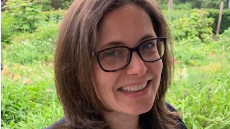 Persoon met bril glimlacht voor een groene tuinachtergrond.