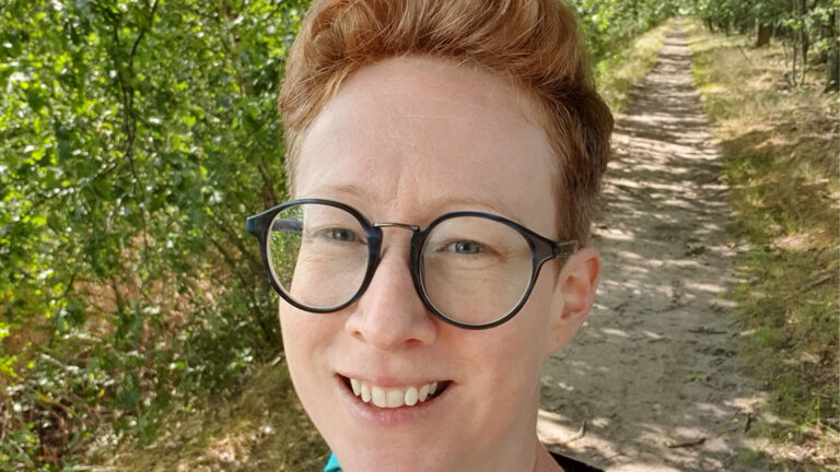 Persoon met bril glimlacht op een bospad met groene bomen op de achtergrond.