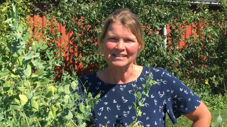 Vrouw in tuin met groene planten, rode muur op achtergrond. Ze glimlacht en draagt een donkerblauwe blouse.