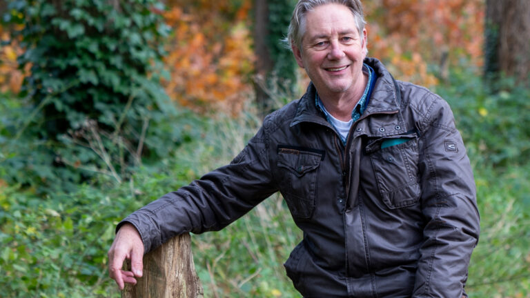 Man in bruine jas leunt op houten hek in bosrijke omgeving met herfstbladeren.