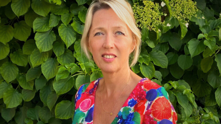 Blonde vrouw in kleurrijke jurk staat voor groene bladeren.