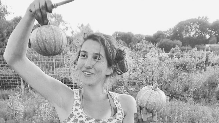 Vrouw in tuin houdt twee pompoenen vast, omringd door planten en hekken.