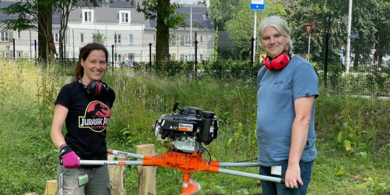 Een man en vrouw bij een machine op een grasveld; ze dragen gehoorbescherming.