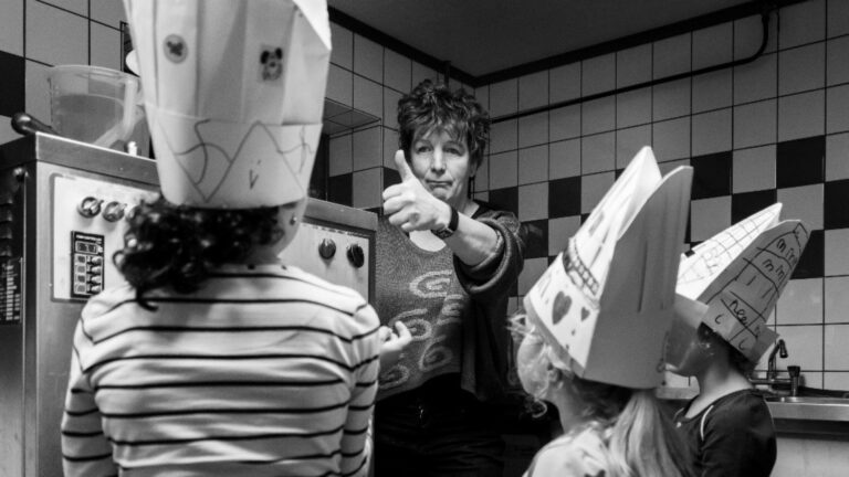 Vrouw geeft duim omhoog naar kinderen met papieren koksmutsen in een keuken.