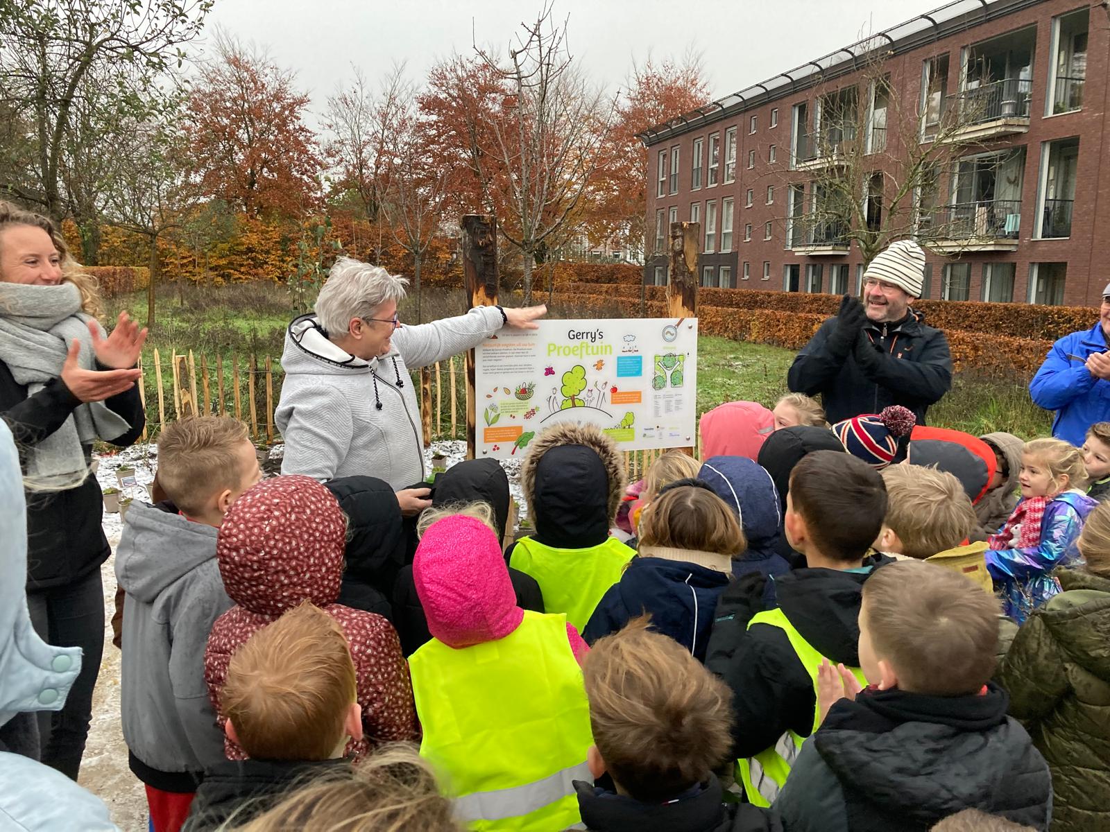 Groep kinderen luistert naar uitleg over een project in een groene omgeving met bomen en gebouwen.