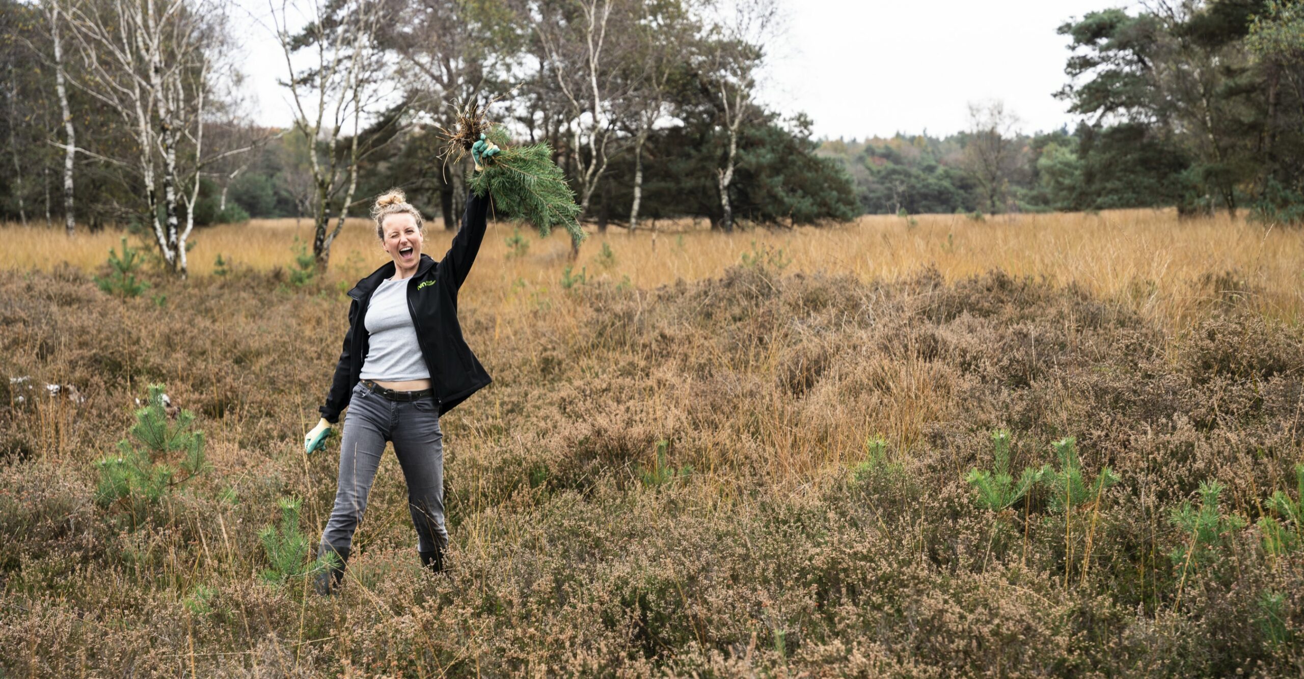 Vrouw met groen in hand in een herfstig natuurgebied, omringd door bomen en bruin gras.