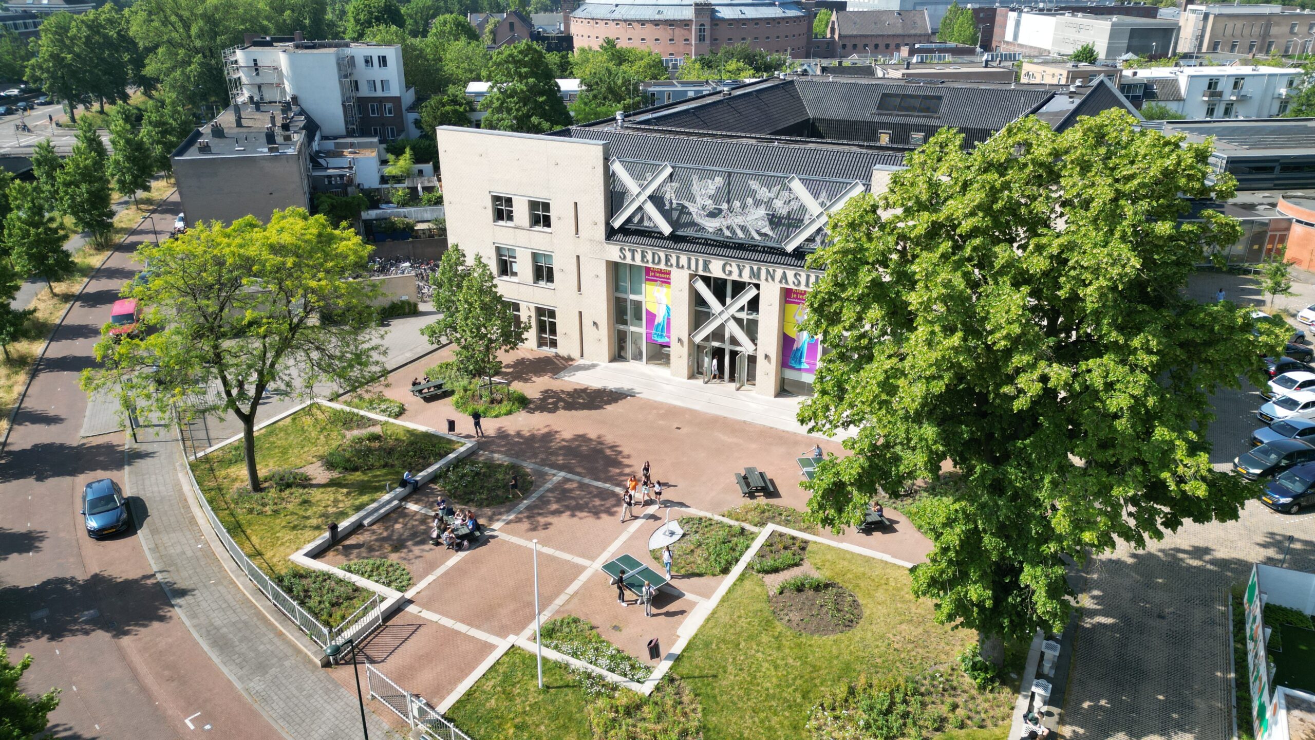Luchtfoto van een schoolgebouw met groene omgeving, pingpongtafel en bankjes, met mensen op het plein.